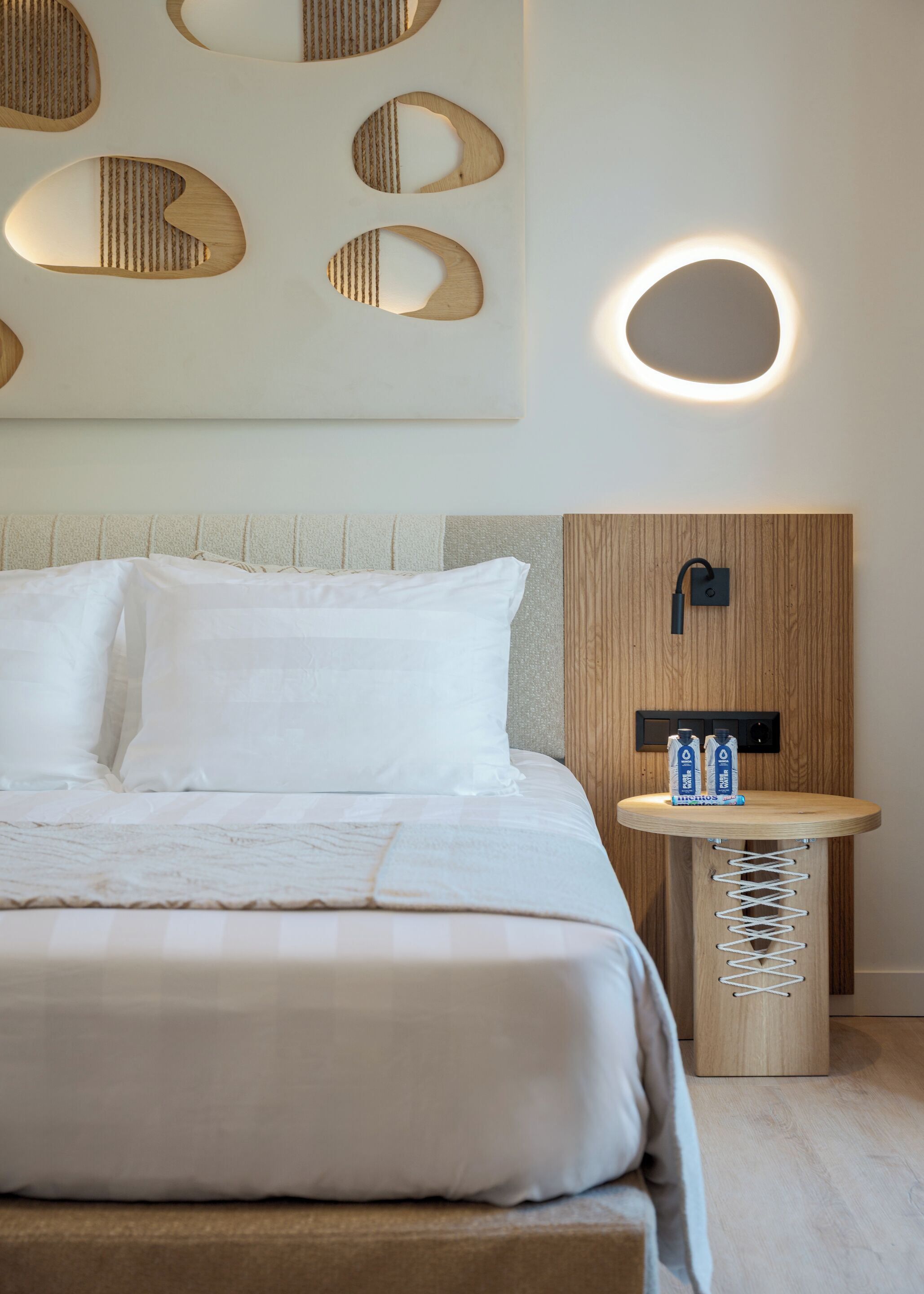 Una habitación de hotel moderna con una cama bien hecha, una mesita de noche redonda con una botella de agua y una lámpara fijada a la pared.