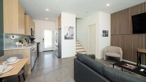 A modern kitchen with wooden cabinets, a dining area with a table and chairs, a gray sofa, and a staircase leading to another level.