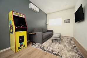 A living room with a gray sofa, a small gray chair, a Pac-Man arcade game, a flat-screen TV, and a patterned rug.