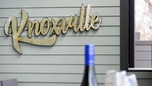 A table with a bottle and two glasses, a metal sign spelling "Knoxville," and a window with blinds.