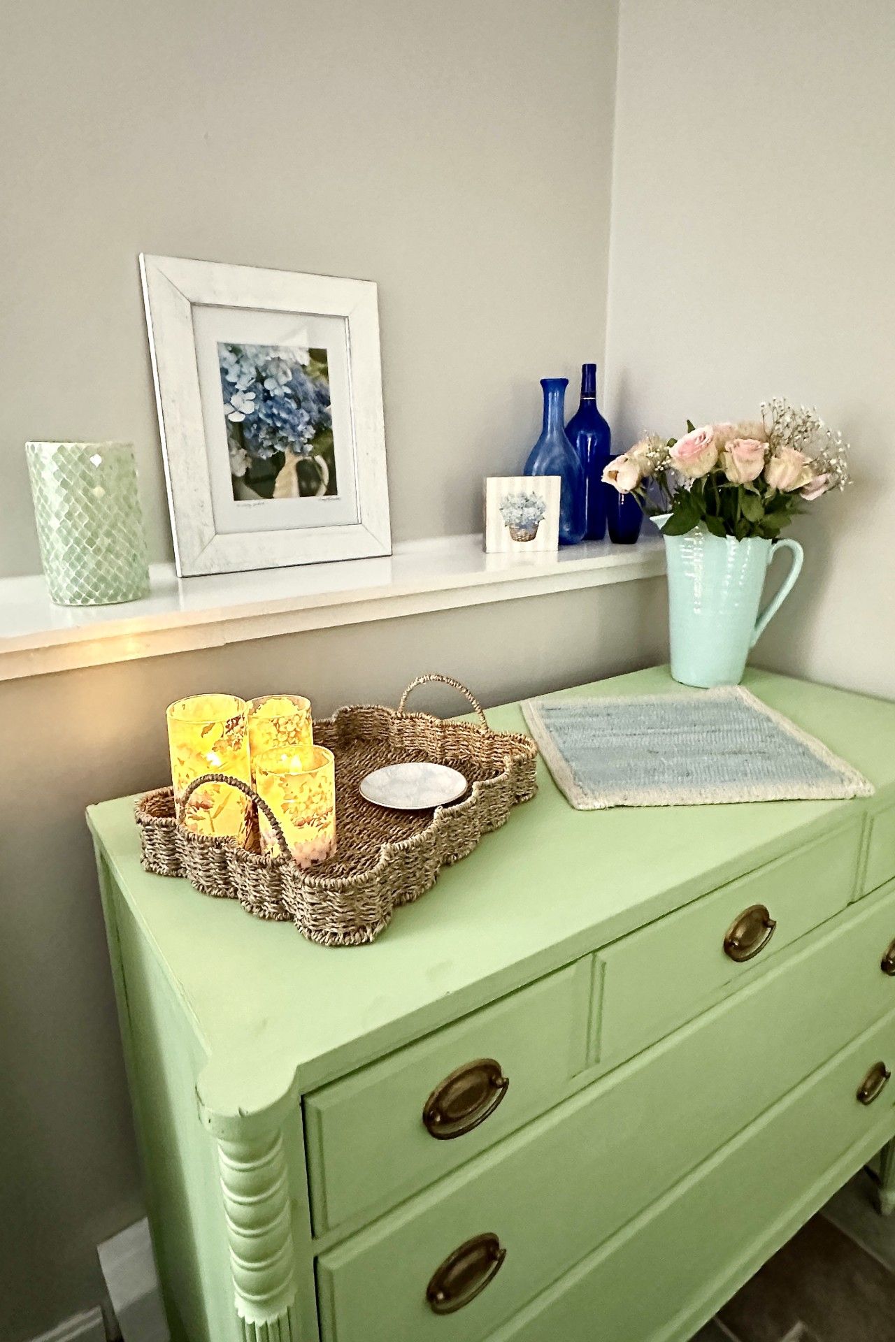 A green dresser with drawers, a woven basket with candles, a framed picture, and decorative vases.