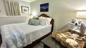 A neatly made bed with a patterned bedspread and pillows, a bedside table with a lamp and framed photos, and a woven basket with starfish on a tiled floor.