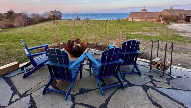A patio with two blue Adirondack chairs facing a large decorative metal bowl, overlooking a grassy area with a view of the ocean and distant buildings.