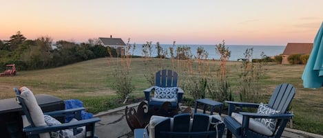 An outdoor seating area with Adirondack chairs and a fire pit, overlooking a body of water during sunset.