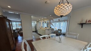 A dining area with a white table and chairs, a hanging light fixture, and a kitchen in the background. - GOLDEN OWL - Town of Owls Head (Owls Head)