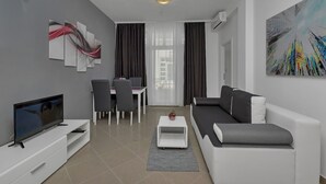 Ein modernes Wohnzimmer mit Flachbildfernseher, einem weißen Sofa, einem Essbereich mit Tisch und Stühlen sowie einem großen abstrakten Gemälde an der Wand.