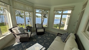 A sunlit room with large windows overlooking a body of water, wicker furniture, and a patterned rug. - BELFAST ON THE BAY - Town of Belfast (Belfast)