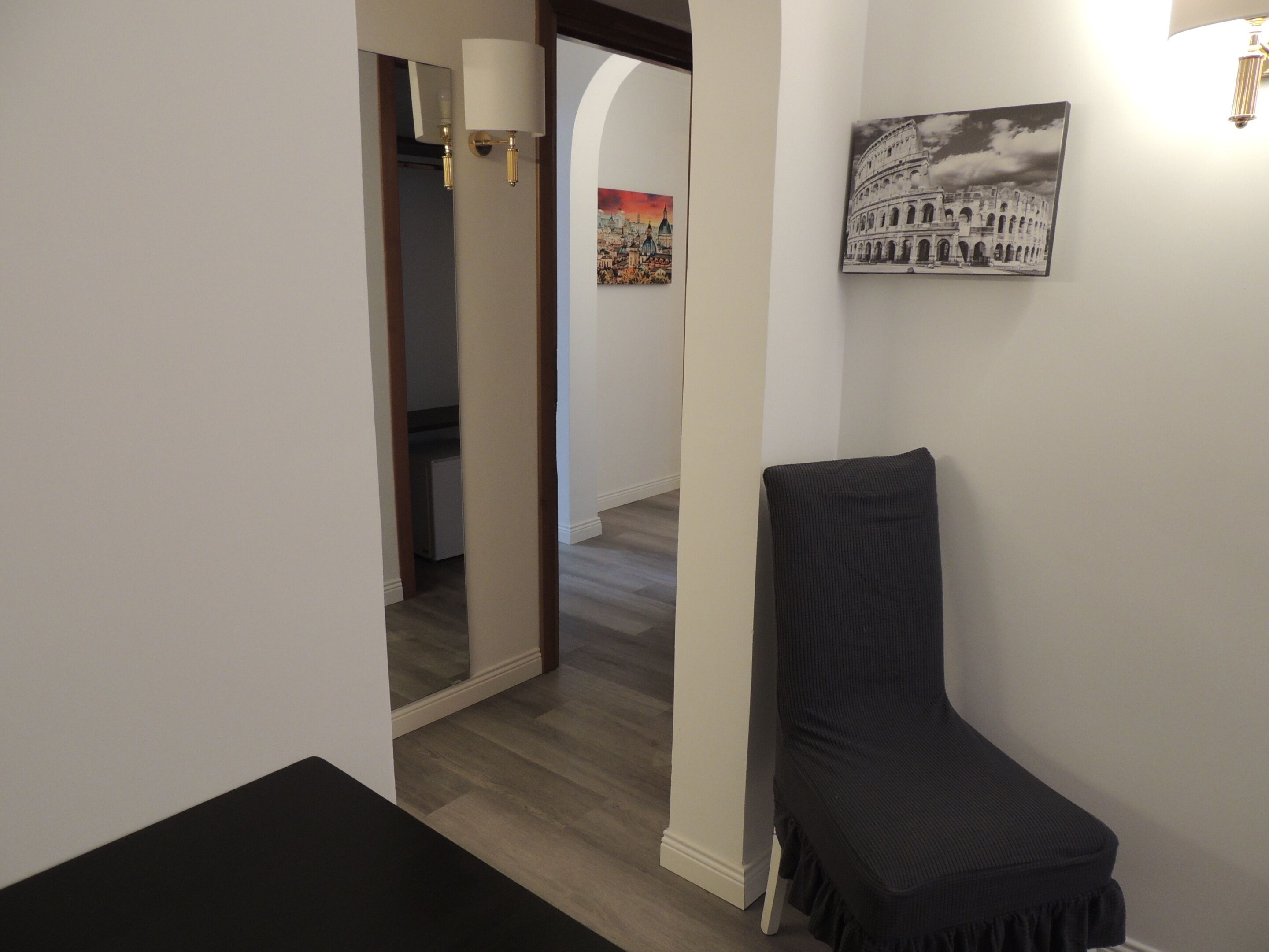 Un corridoio stretto con una sedia nera, un muro bianco con un quadro che raffigura il Colosseo e una porta che dà su un'altra stanza.