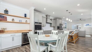 A modern kitchen with white cabinetry, a central island with chairs, and a dining area with a round table. - High Tide 3BR Townhome North OC (Ocean City)