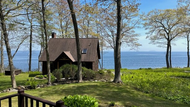 A cabin with a sloped roof, surrounded by trees and a grassy area, overlooking a body of water.