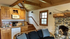 A cozy kitchen with wooden cabinets, a microwave, and a coffee maker. There's a wicker sofa with cushions and a stone fireplace with logs.