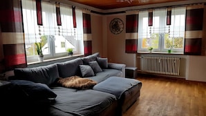Un salon chaleureux avec un canapé gris, un plaid en fausse fourrure et un radiateur. Il y a des fenêtres avec des rideaux à rayures et une horloge au mur.