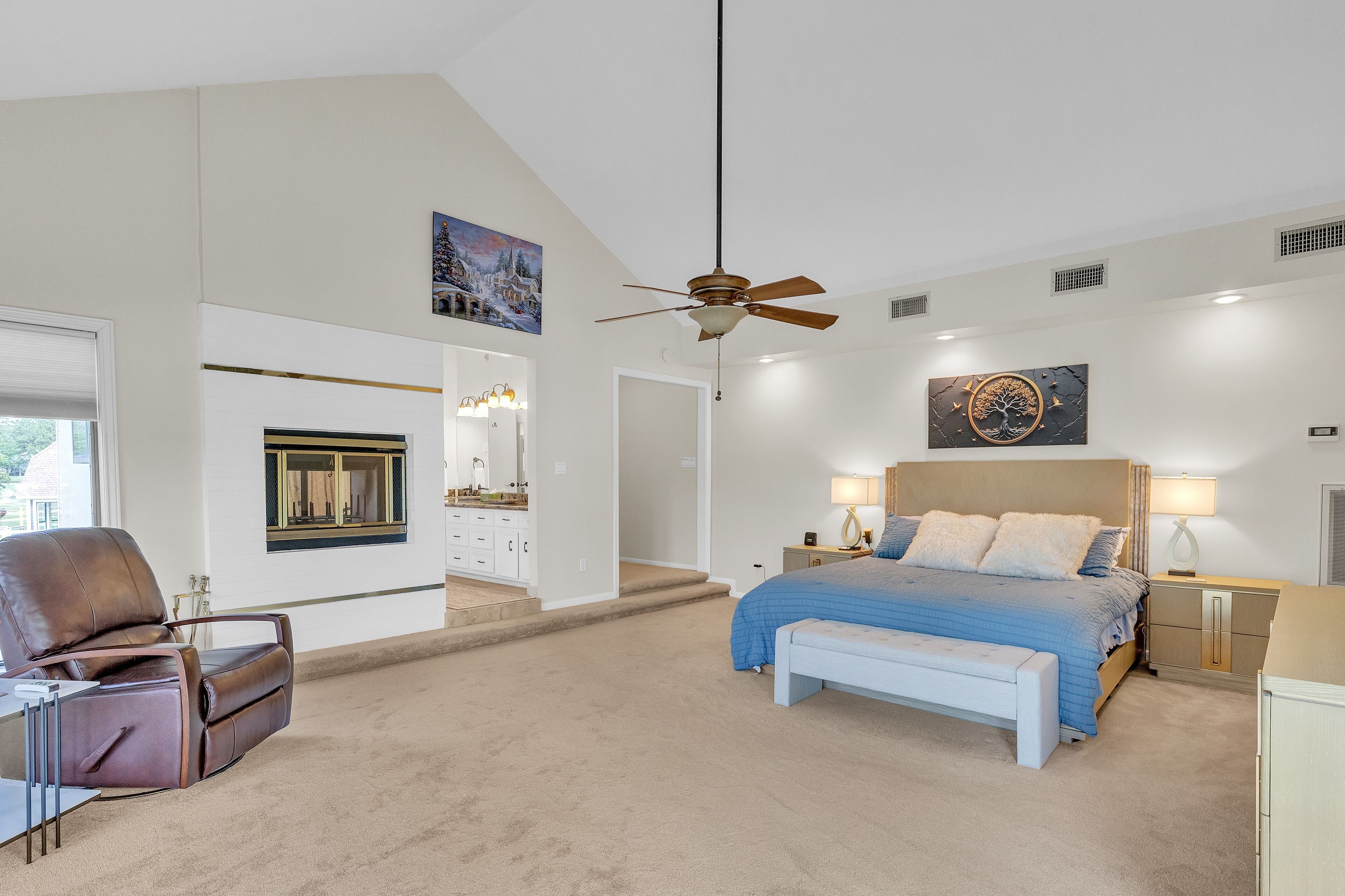 A bedroom with a bed, a chair, a bench, a ceiling fan, and artwork on the walls.
