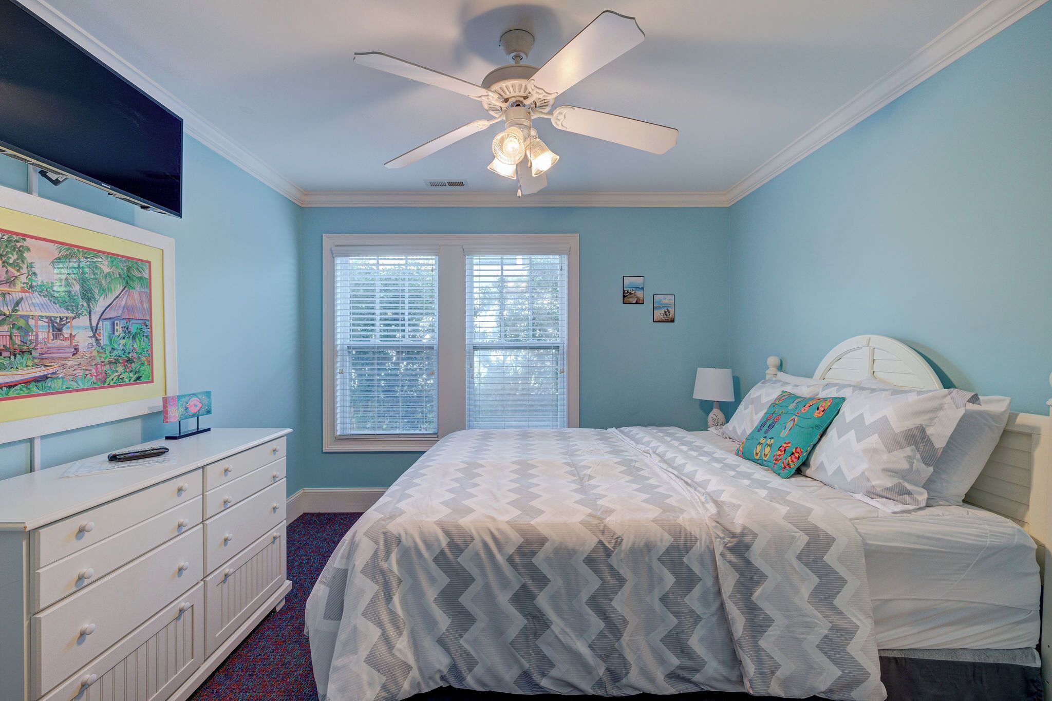 Un dormitorio con cama, ventilador de techo, cómoda, televisor y ventana con persianas.