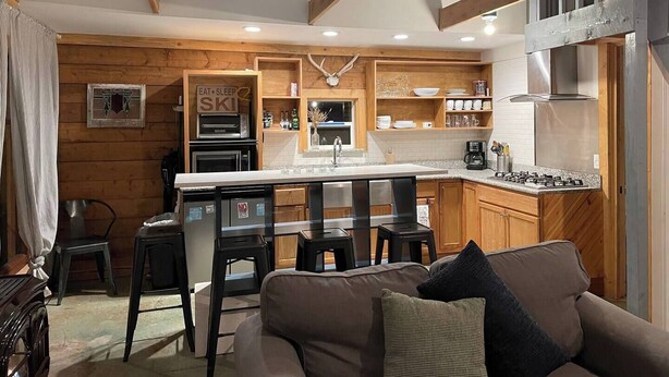 A modern kitchen with wooden cabinetry, a central island, and a dining area with bar stools. - Walk to Downtown Creede | Pet-Friendly | Mountain Views | Fenced Yard (Creede)