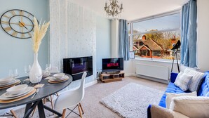 Una sala de estar moderna con comedor, un ventanal con vistas a casas, un televisor mostrando 'NETFLIX' y una araña.