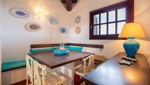 A dining area with a wooden table, white wicker chairs, a blue bowl on the table, and decorative plates on the wall.