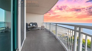 A balcony with a view of the ocean at sunset, a table with chairs, and a railing.