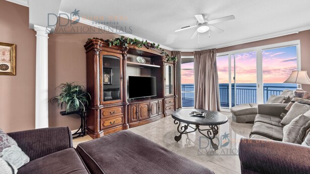 A spacious living room with a large TV, comfortable seating, a ceiling fan, and a balcony with a scenic view.