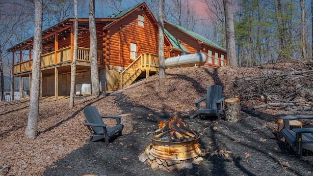 A log cabin with a deck and a fire pit surrounded by Adirondack chairs.