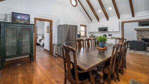 A spacious dining area with a long wooden table and chairs, a stone fireplace, and a kitchen with white cabinets and wooden beams.