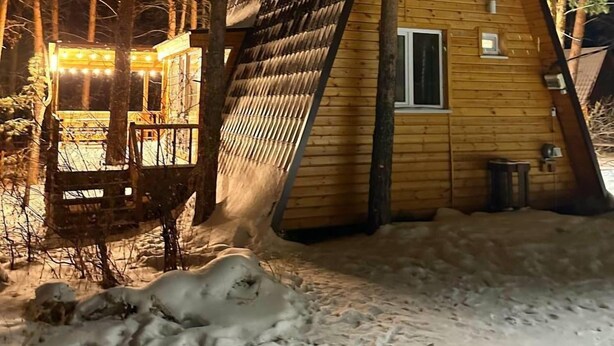A triangular wooden cabin with a sloped roof, surrounded by trees and covered in snow.