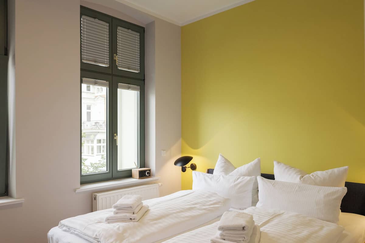 A hotel room with two beds, a window with blinds, a radiator, and a lamp.