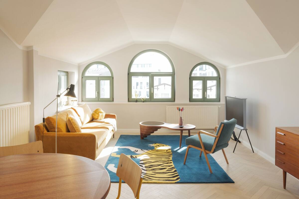 A spacious loft apartment with a high ceiling, a cozy living area with a sofa and armchairs, a dining table, and a large window with arched tops.