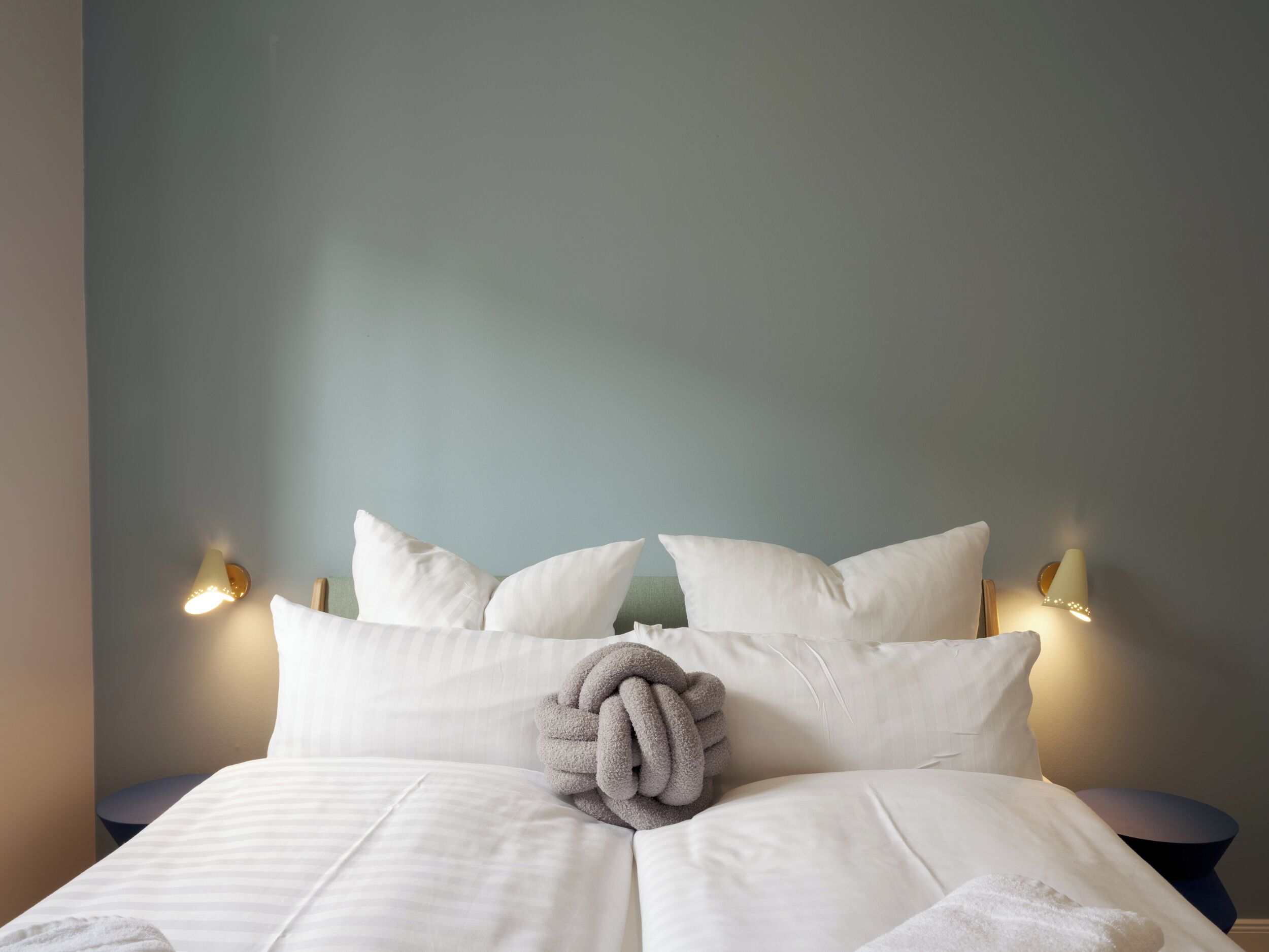 Ein ordentlich gemachtes Bett mit weißer Bettwäsche und einer grauen Strickdecke, zwei Wandlampen und eine schlichte Wand.