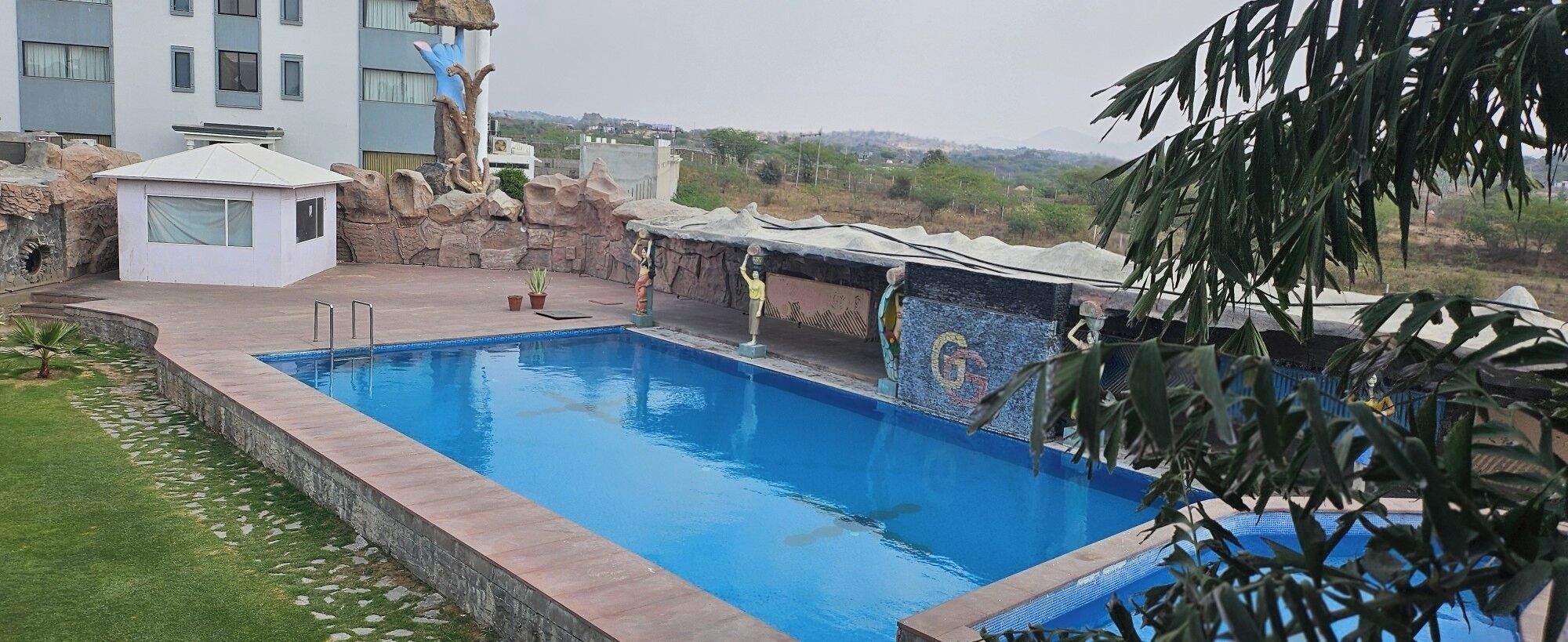 Una piscina con borde de piedra, un pequeño jardín y vista a un edificio al fondo.