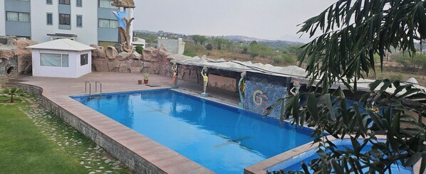 Una piscina con borde de piedra, un pequeño jardín y vista a un edificio al fondo.