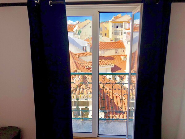 A view through a window with a metal balcony railing, a curtain partially drawn, and buildings with red-tiled roofs in the background. - Room-3lisbon Downtown Private Bedroom Ri (Lisbon)