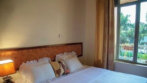 A neatly made bed with white linens and decorative pillows, a bedside lamp, and a window with a view of greenery outside. - HOTEL GREENWICH (Kigali)