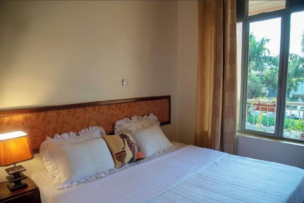 A neatly made bed with white linens and decorative pillows, a bedside lamp, and a window with a view of greenery outside.