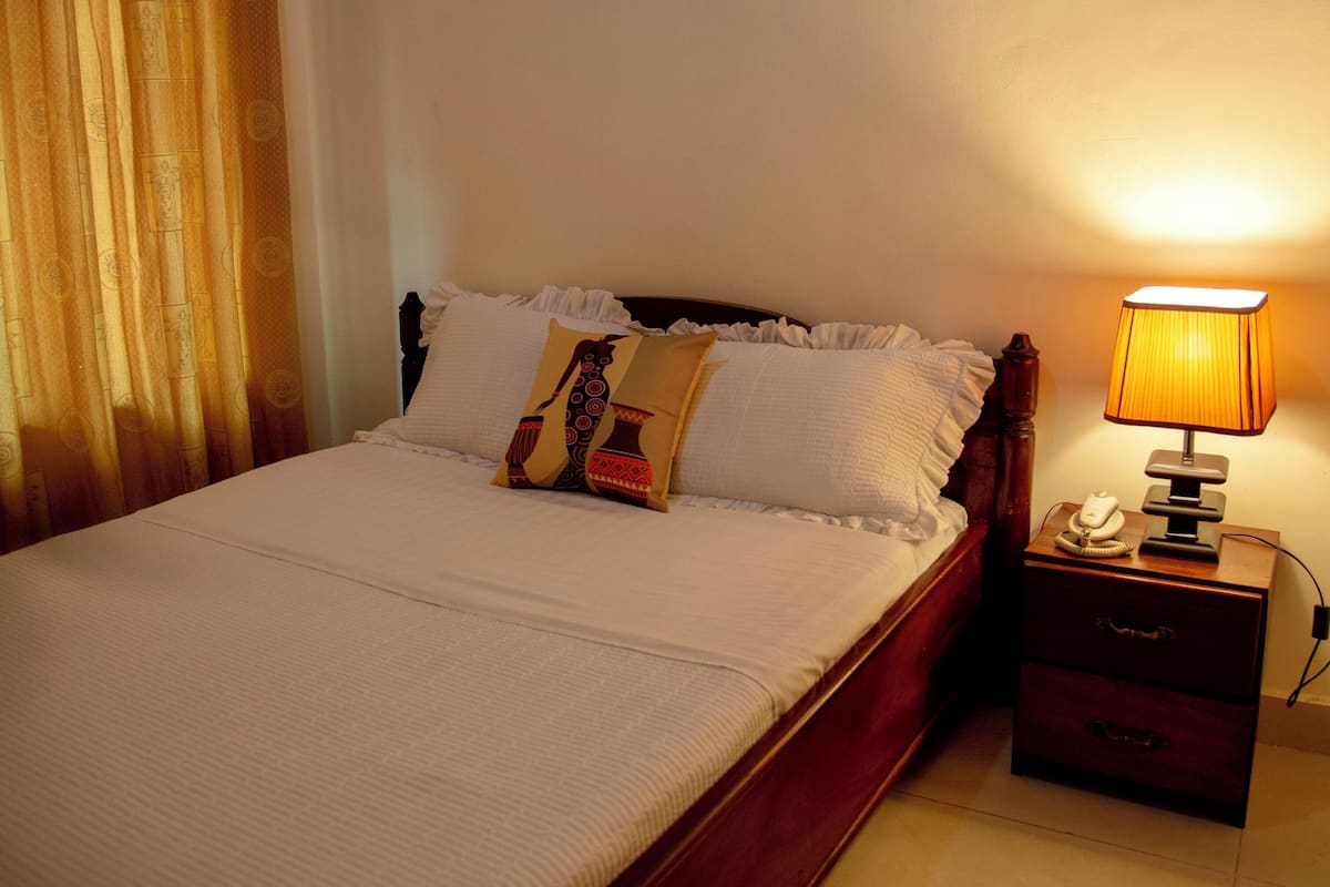 A neatly made bed with white bedding and decorative pillows, a bedside lamp, and a wooden nightstand with a phone and a drawer.