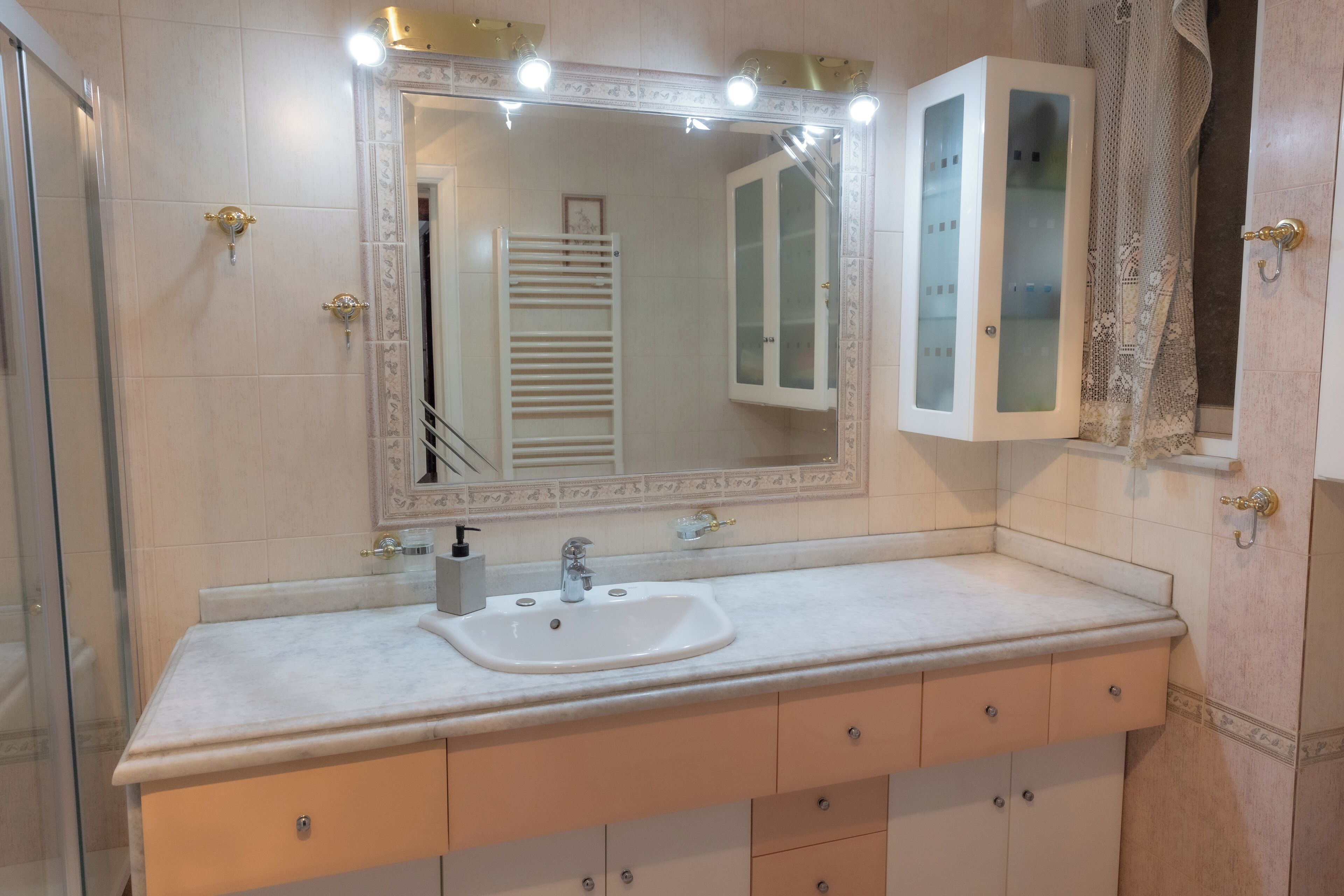Een badkamer met een wit wastafelmeubel, een grote spiegel met verlichting, een glazen kast en een douchehoek.