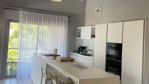 A modern kitchen with a central island, bar stools, a large window with sheer curtains, and a hanging light fixture.
