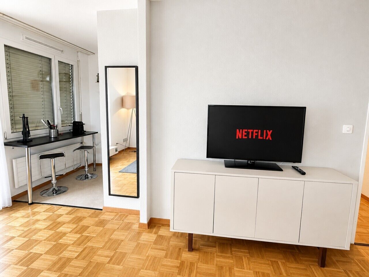 A modern living room with a flat-screen TV displaying 'NETFLIX,' a white cabinet, and a kitchen area with a bar stool and a coffee machine.