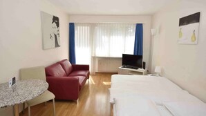 A hotel room with a bed, a red sofa, a TV, a small table, and a window with curtains. - Cozy studio in the city center - Crown 34 (Zürich)