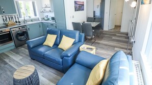 A modern living room with a blue sofa, a round wooden coffee table, and a dining area with a table and chairs.