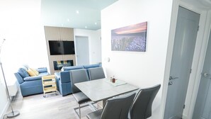 A modern living room with a sofa, a dining table with chairs, a TV mounted on a wall, and a door.