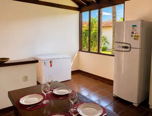 A compact kitchen with a dining table set for two, a white refrigerator, and a window with a view of greenery. - Bz11 Geribá Casa Piscina sauna 6 quartos (Buzios)