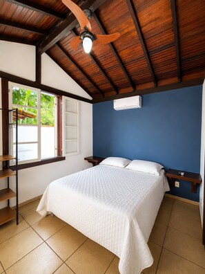 Una habitación con cama, ventilador de techo, ventana con vista a follaje y aire acondicionado de pared.