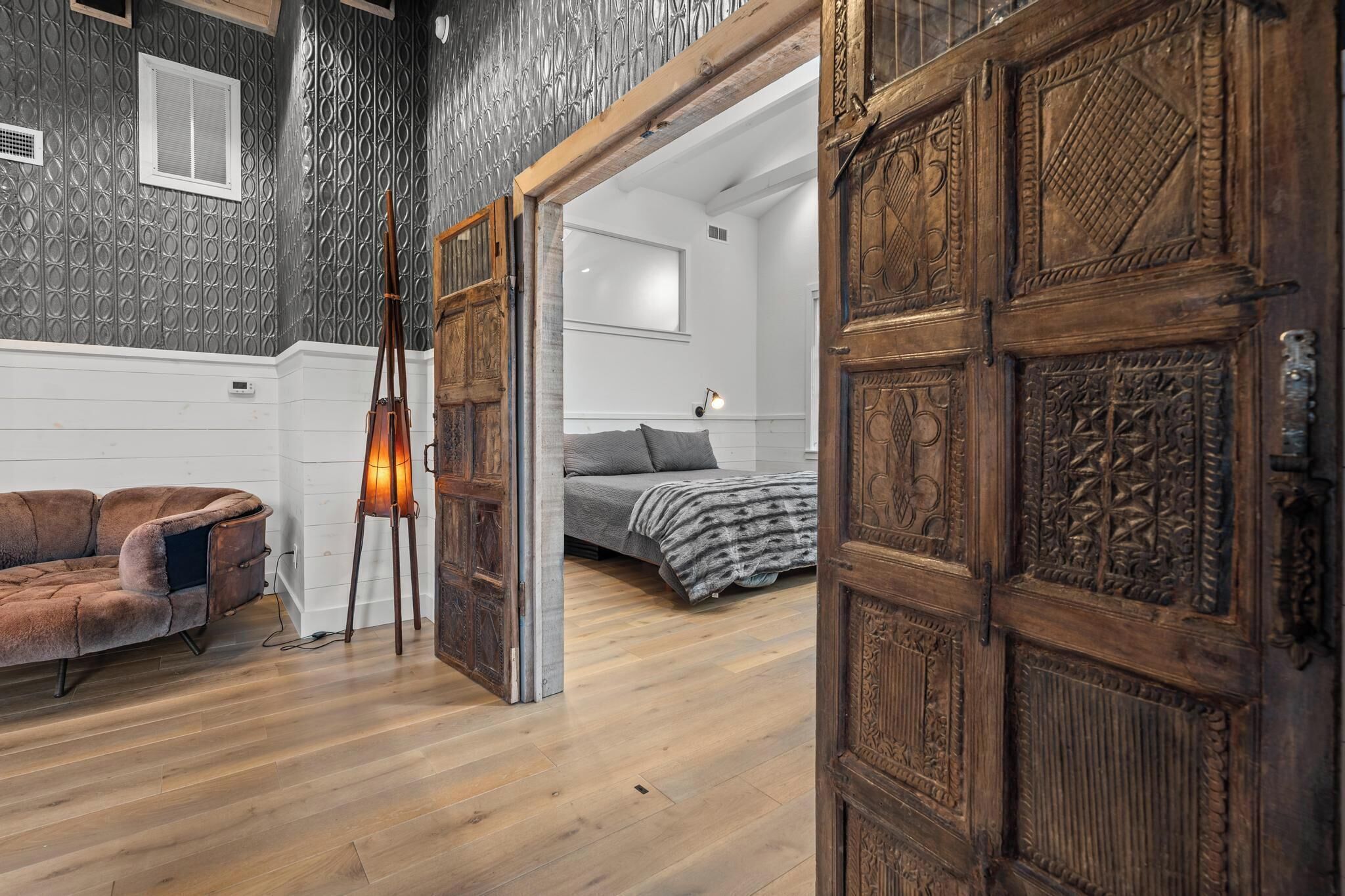 A room with a wooden floor, a brown sofa, a bed with a gray blanket, and a wooden door with intricate carvings.