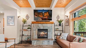 A cozy living room with a stone fireplace, wooden ceiling beams, and a large window overlooking a snowy landscape.