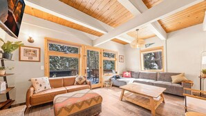 A cozy living room with a leather sofa, a wooden coffee table, and a grey couch. Large windows offer views of snow-covered trees.