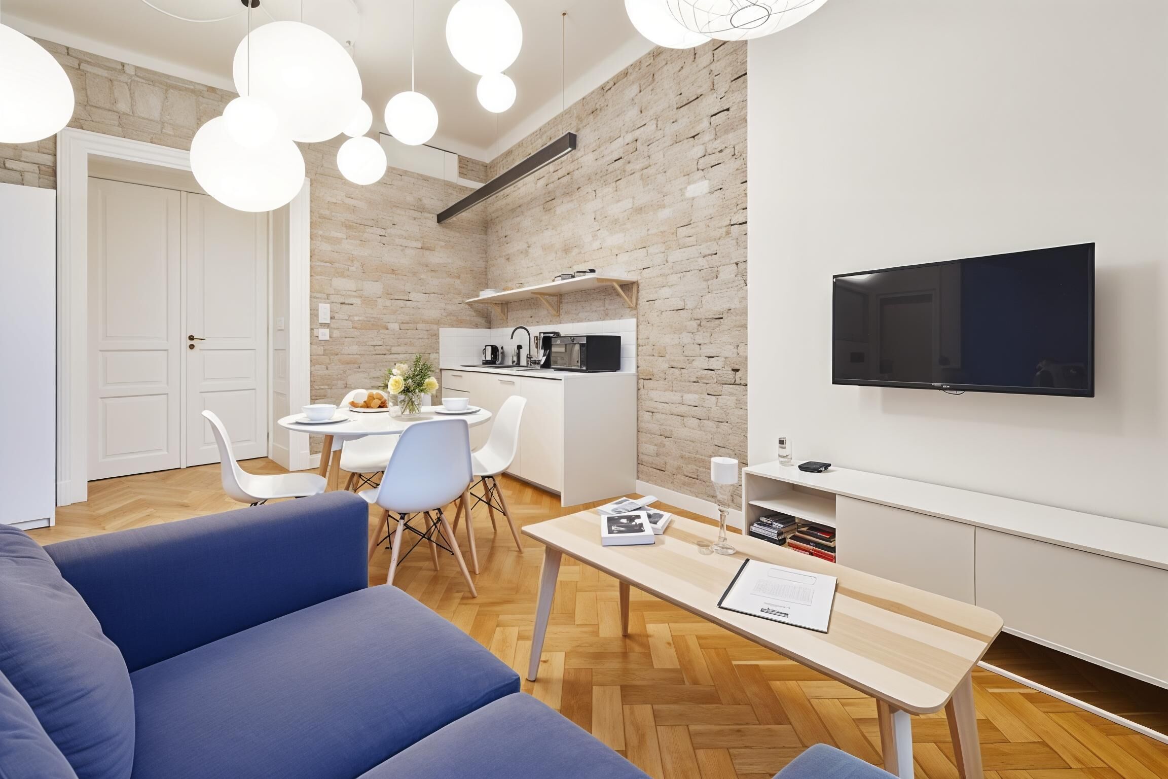 A modern living room with a blue sofa, a dining area with a white table and chairs, a kitchen with a brick wall, and a mounted TV.