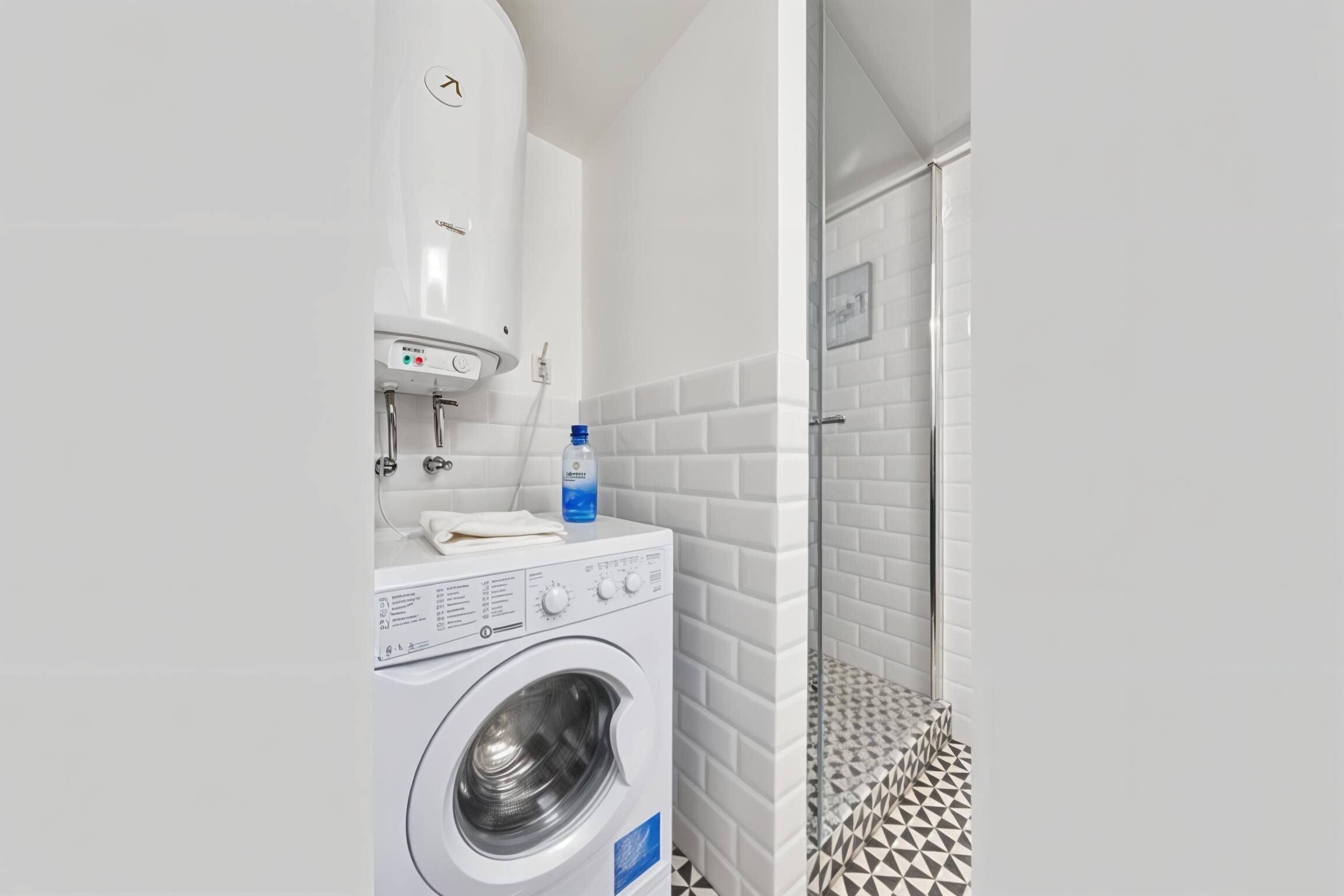 Ein kleiner Waschbereich mit einer Waschmaschine, einem Durchlauferhitzer und einer Dusche mit fliesenbelagtem Boden in geometrischem Muster.
