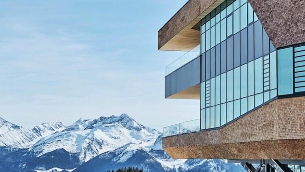 Ein modernes Gebäude mit Holzfassade und großen Fenstern, vor einer Bergkulisse gelegen.