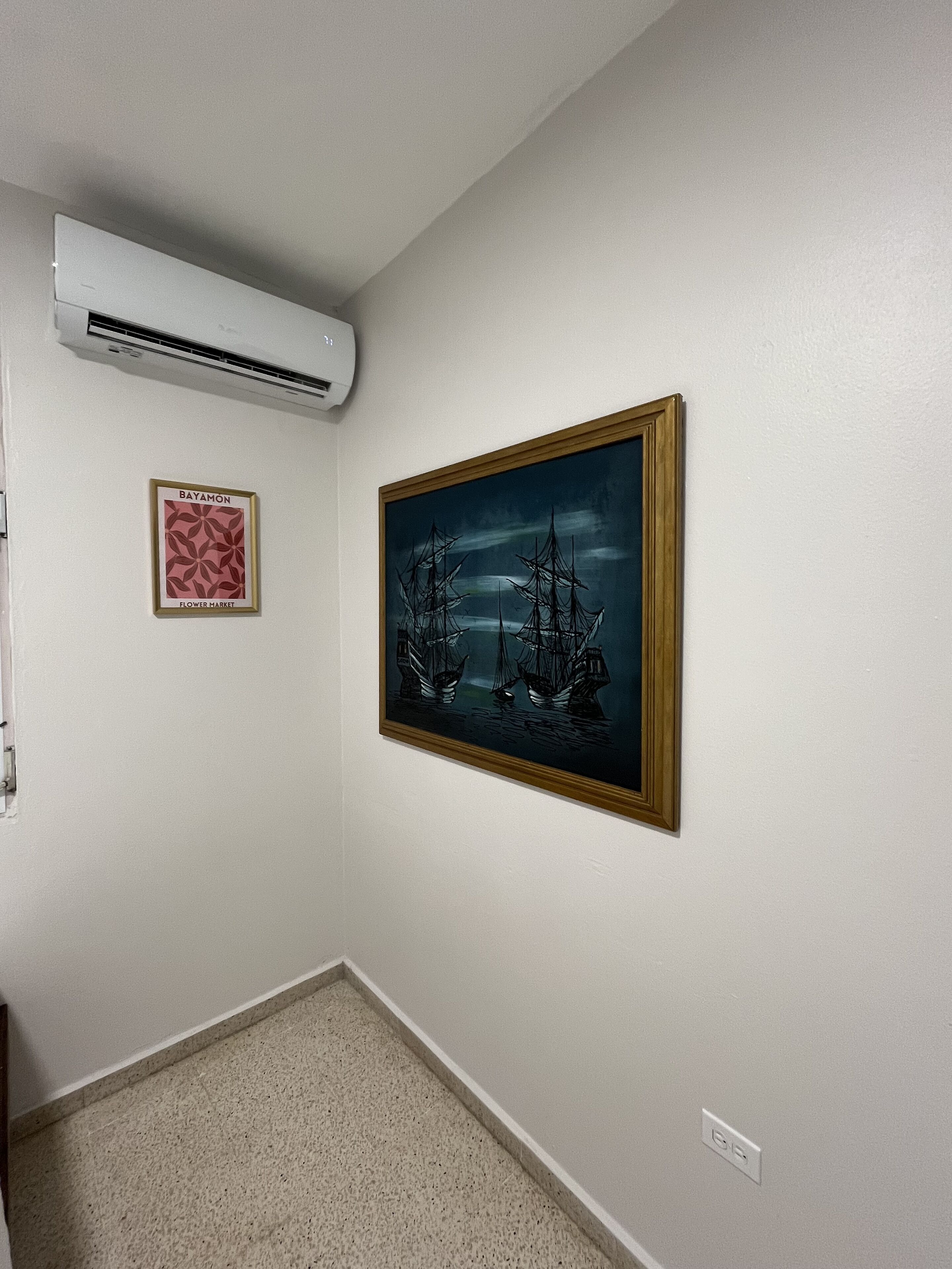 Un couloir orné d’un tableau encadré représentant des navires, d’un climatiseur fixé au mur et d’un petit panneau encadré.
